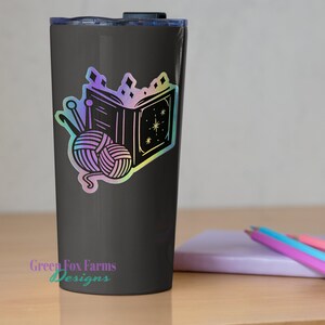 May include: A black tumbler with a holographic sticker of a book, yarn, and knitting needles. The sticker has a rainbow iridescent finish. The tumbler has the text "Green Fox Farms Designs" on the bottom.