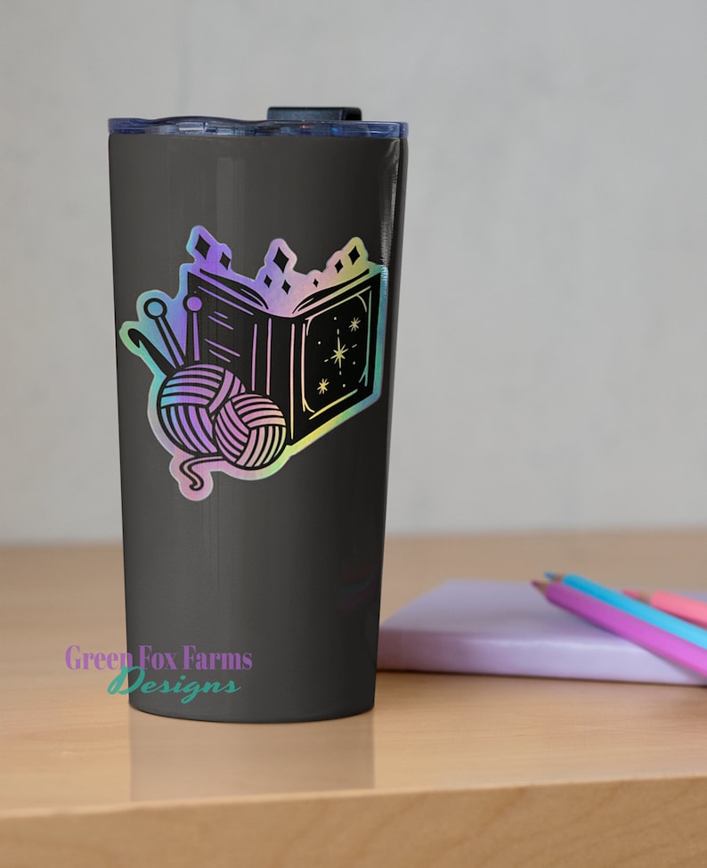 May include: A black tumbler with a holographic sticker of a book, yarn, and knitting needles. The sticker has a rainbow iridescent finish. The tumbler has the text "Green Fox Farms Designs" on the bottom.