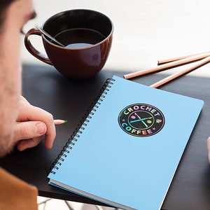 May include: A blue spiral notebook with a black and rainbow holographic sticker that says "Crochet & Coffee" with a pair of crossed crochet needles and coffee beans.