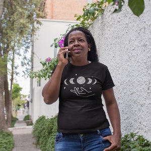 May include: A black t-shirt with a white design of a hand holding a moon with stars. The moon is in different phases.