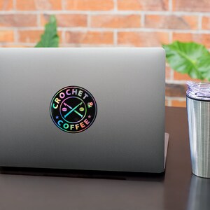 May include: A silver laptop with a black and iridescent sticker that says "Crochet & Coffee" with a pair of crossed knitting needles and coffee beans. A silver travel mug with a purple lid is to the right of the laptop.