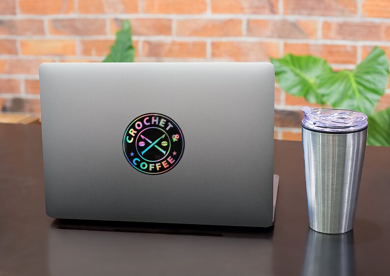 May include: A silver laptop with a black and iridescent sticker that says "Crochet & Coffee" with a pair of crossed knitting needles and coffee beans. A silver travel mug with a purple lid is to the right of the laptop.