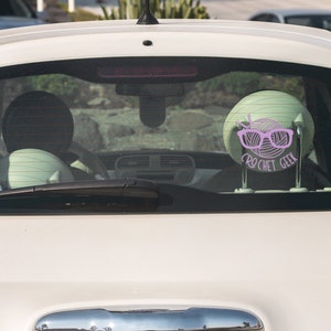 May include: A white car with a pink crochet geek sticker on the back window. The sticker features a ball of yarn with knitting needles and glasses.
