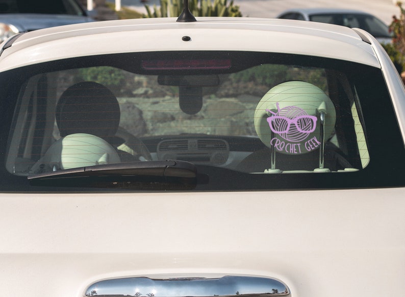 May include: A white car with a pink crochet geek sticker on the back window. The sticker features a ball of yarn with knitting needles and glasses.