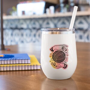 May include: White stainless steel wine tumbler with a colorful sticker of two hands holding a ball of yarn. The sticker has a pink, orange, and yellow gradient.