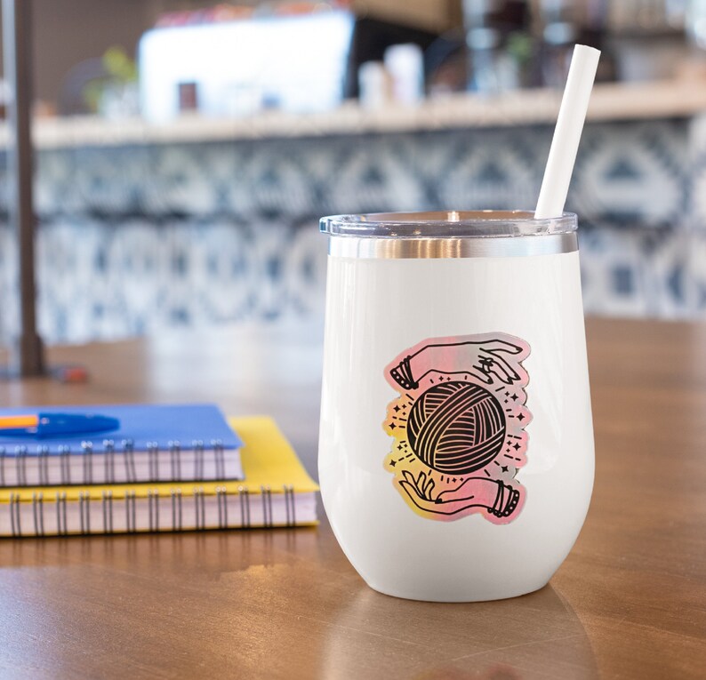May include: White stainless steel wine tumbler with a colorful sticker of two hands holding a ball of yarn. The sticker has a pink, orange, and yellow gradient.