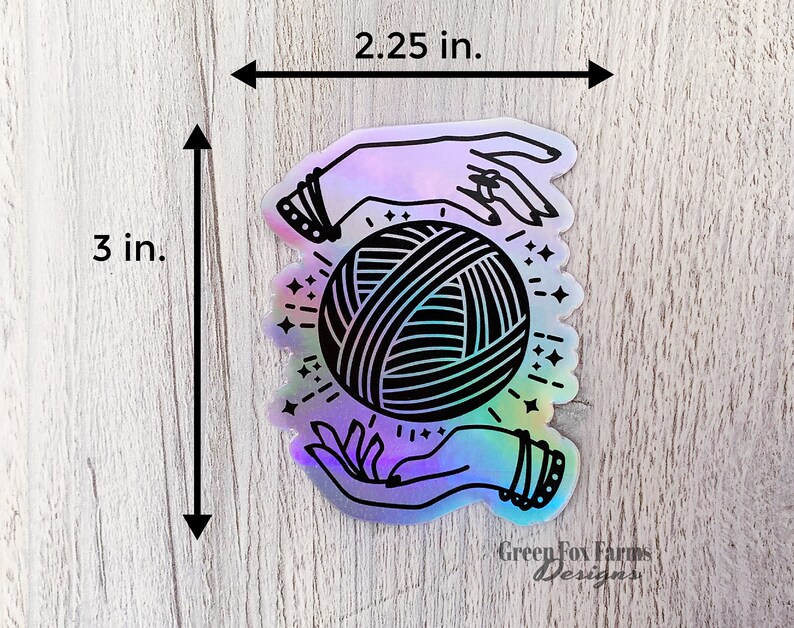 May include: A holographic sticker featuring a pair of hands holding a ball of yarn with a black and white design. The sticker is 3 inches tall and 2.25 inches wide. The sticker has the text "Green Fox Farms Designs" at the bottom.
