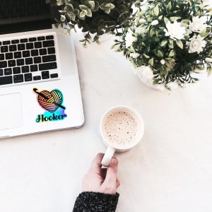 May include: A laptop computer with a sticker of a heart-shaped yarn ball and the word "Hooker" on it. A cup of coffee sits on a white surface next to the laptop.
