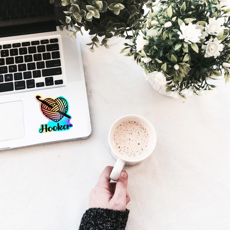 May include: A laptop computer with a sticker of a heart-shaped yarn ball and the word "Hooker" on it. A cup of coffee sits on a white surface next to the laptop.