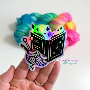 May include: A holographic sticker featuring a black and white illustration of an open book with a ball of yarn and knitting needles. The book has stars and diamonds on it. The sticker has a rainbow iridescent finish. The text "Green Fox Farms Designs" is visible at the bottom of the sticker.