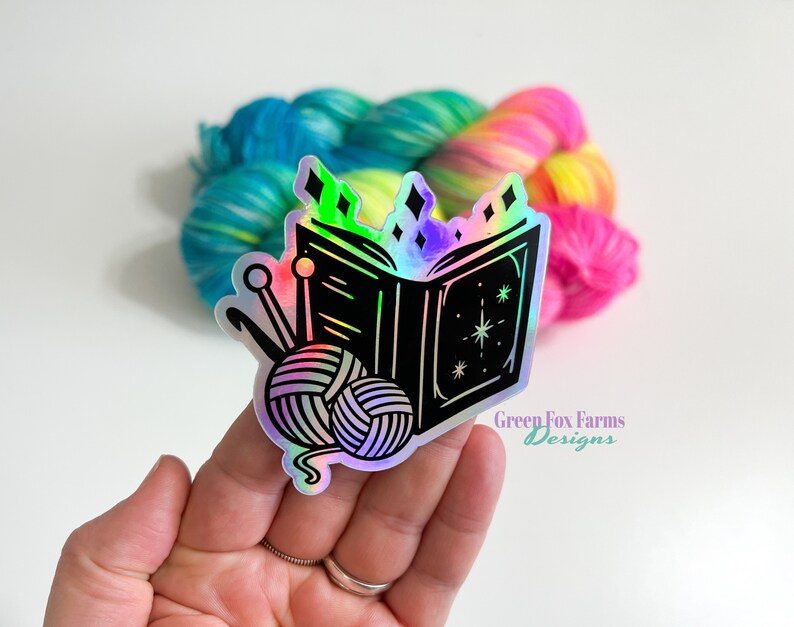 May include: A holographic sticker featuring a black and white illustration of an open book with a ball of yarn and knitting needles. The book has stars and diamonds on it. The sticker has a rainbow iridescent finish. The text "Green Fox Farms Designs" is visible at the bottom of the sticker.