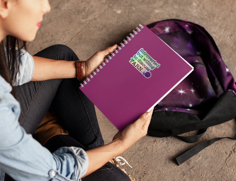 May include: A purple spiral bound notebook with a sticker that says "Introverted but willing to discuss YARN."
