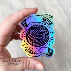 May include: A holographic sticker featuring two hands holding a ball of yarn. The yarn is black and white and the hands are iridescent.
