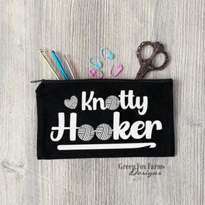May include: Black zippered pouch with the text "Knotty Hooker" in white with a heart and yarn ball design. The pouch is on a woodgrain surface with crochet hooks and stitch markers.