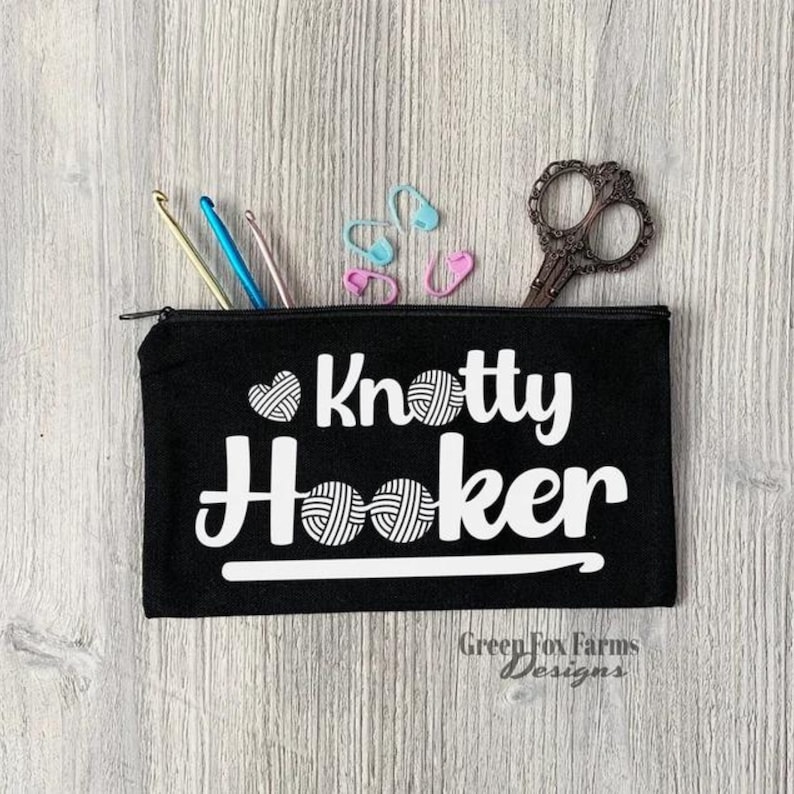 May include: Black zippered pouch with the text "Knotty Hooker" in white with a heart and yarn ball design. The pouch is on a woodgrain surface with crochet hooks and stitch markers.