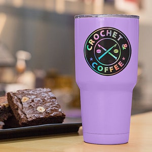 May include: A purple stainless steel tumbler with a black and iridescent sticker that says "Crochet & Coffee" with a crossed crochet hook and coffee bean design.