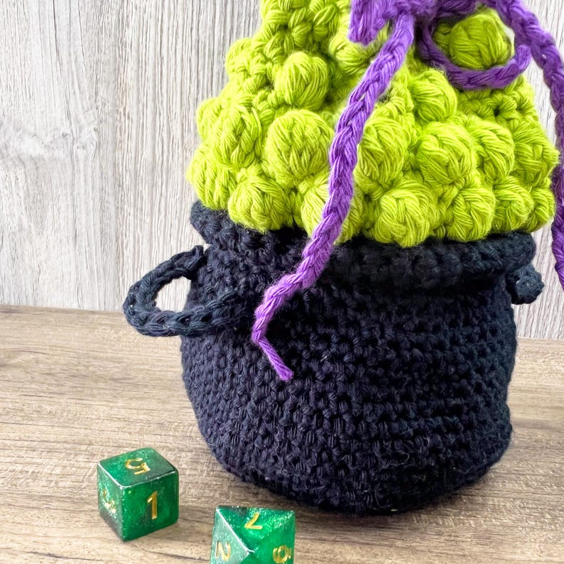 May include: A handmade crochet cauldron in black with a lime green, textured top. The cauldron has two small handles and a purple drawstring. Two green dice with white numbers are in the foreground. A unique Halloween or fantasy-themed item.
