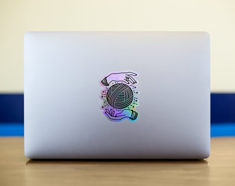 May include: A holographic sticker with a black and white illustration of two hands holding a ball of yarn. The sticker has a rainbow iridescent finish.