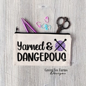 May include: A white canvas zippered pouch with the text "Yarned & Dangerous" in black lettering. A purple ball of yarn with two knitting needles crossed over it is to the right of the text. The pouch is partially open and contains two pencils, a pair of scissors, and several stitch markers.