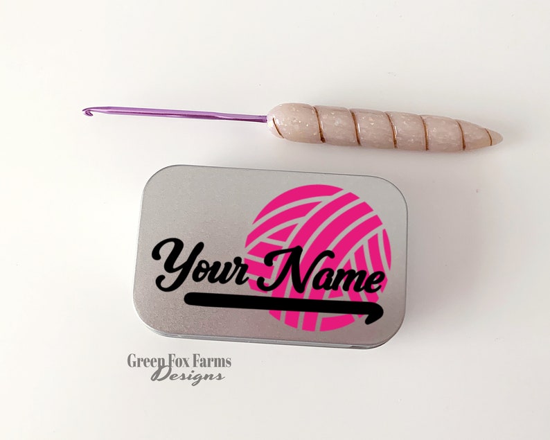 May include: A silver tin with a pink yarn ball and a black crochet hook graphic. The text "Your Name" is printed in black on the tin. A purple crochet hook is also pictured.