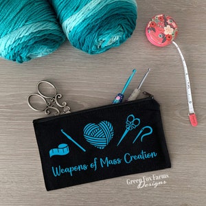 May include: Black canvas zippered pouch with the text "Weapons of Mass Creation" and a blue graphic of yarn, scissors, a crochet hook, and a tape measure. The pouch also has the text "Green Fox Farms Designs" in white.