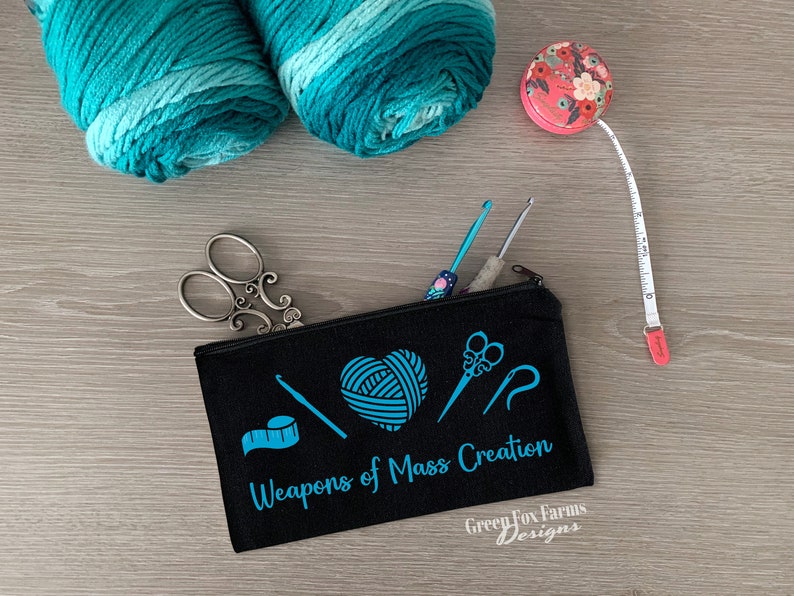 May include: Black canvas zippered pouch with the text "Weapons of Mass Creation" and a blue graphic of yarn, scissors, a crochet hook, and a tape measure. The pouch also has the text "Green Fox Farms Designs" in white.