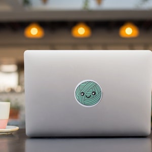 May include: A silver laptop computer with a green yarn ball sticker on the lid. The yarn ball has a cute cartoon face with eyes and a smile.