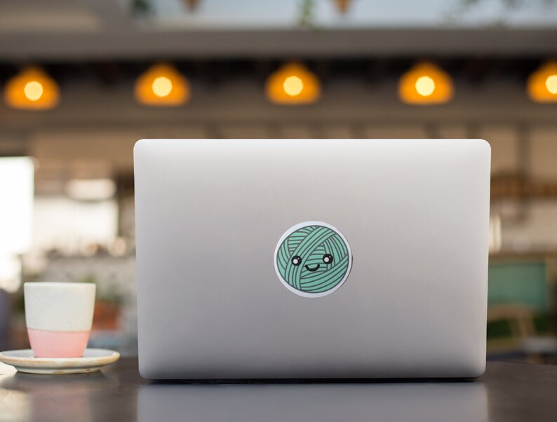May include: A silver laptop computer with a green yarn ball sticker on the lid. The yarn ball has a cute cartoon face with eyes and a smile.