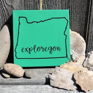 May include: A square canvas featuring a bright green background with a black outline of the state of Oregon. The word exploregon is printed in black cursive script across the center of the state outline, positioned on a wooden surface.