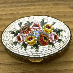 May include: Oval-shaped trinket box with a mosaic floral design. The lid features a mosaic of colorful flowers, including red, pink, and yellow roses, set against a white background. The box has a brass-colored metal frame.