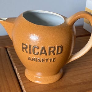 Pode incluir: Um jarro de cerâmica castanho com um corpo arredondado, um bico e uma pega. As palavras "RICARD ANISSETTE" estão em relevo em letras castanhas na frente. O interior é azul claro.