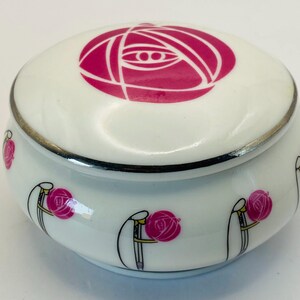 May include: A white ceramic trinket box with a pink rose design on the lid. The box has a silver rim and is decorated with pink roses and black line art. The box is oval-shaped.