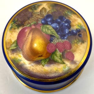 May include: A round, blue trinket box with a gold rim and a painted fruit design on the lid. The artwork features a pear, grapes, raspberries, and leaves. The box is a decorative item.