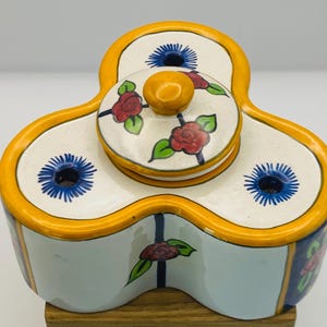 Può includere: Un contenitore in ceramica a tre lobi con coperchio. Il contenitore è dipinto con una base bianca, finiture gialle e disegni floreali blu e rossi. Il coperchio ha un piccolo pomello giallo. Il contenitore viene probabilmente utilizzato per conservare spezie o altri piccoli oggetti.