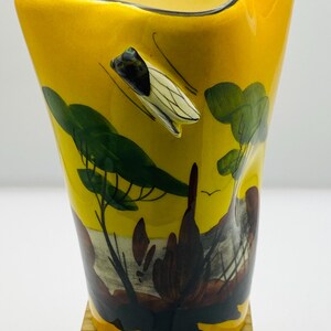 May include: Yellow ceramic vase with a black rim and a unique insect-shaped handle. The vase features a landscape design with green trees and a flying insect. The vase is set on a wooden base.
