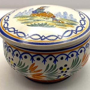May include: A white ceramic lidded jar with a blue, orange, and green floral design. The lid features a painted scene of a person sitting on a mushroom.