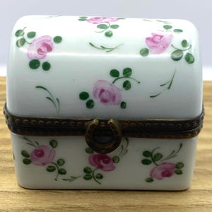 May include: A small, white porcelain trinket box with a hinged lid. The box is decorated with pink roses and green leaves. It has a bronze-coloured metal trim and a horseshoe-shaped clasp.