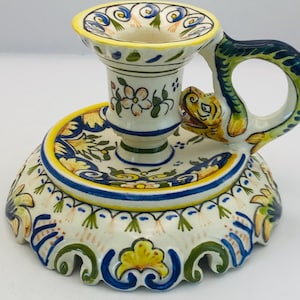 May include: A hand-painted ceramic candlestick holder with a floral and serpent design. The base and cup are white with blue, yellow, and green accents. The handle is a serpent with a yellow head and green body.