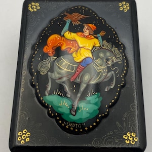 Pode incluir: Caixa lacada preta com uma cena pintada à mão de uma figura a cavalo, segurando um pássaro. A figura usa uma túnica amarela e botas vermelhas. A caixa tem detalhes dourados e uma borda decorativa.