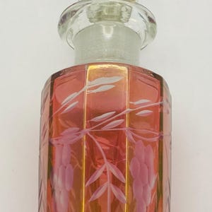 May include: A vintage perfume bottle with a faceted glass stopper. The bottle is a reddish-pink colour with etched floral designs. The bottle has a clear glass neck and stopper.