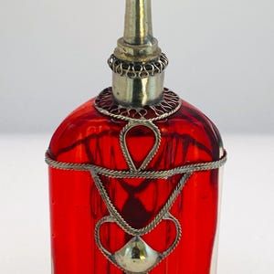 May include: A red glass perfume bottle with a silver metal filigree design. The bottle has a long, slender silver stopper and is adorned with intricate metalwork around the neck and body. The bottle's shape is rounded, with a flat base.