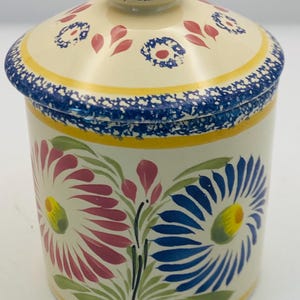 May include: A ceramic jar with a lid, featuring a floral design. The jar is cream-colored with blue, yellow, pink, and green painted flowers. The lid and rim have blue speckled patterns. The jar is approximately 5 inches tall.