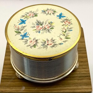 Puede incluir: Una caja de joyas redonda y vintage con tapa crema con diseño floral y mariposas. La caja tiene un borde dorado y un cuerpo plateado y acanalado. Se apoya sobre pequeños pies plateados.