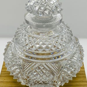May include: A clear, cut-glass perfume bottle with a faceted, diamond-patterned design. The bottle has a round base and a matching stopper. The intricate cuts create a sparkling effect, reflecting light. The bottle sits on a wooden surface.