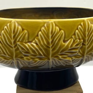 Op de afbeelding: Een gele keramische kom met een reliëf van bladeren. De kom heeft een donkerblauwe voet en staat op een houten oppervlak.