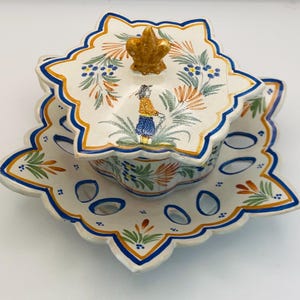 Peut inclure: Un présentoir à œufs en céramique décoratif avec un couvercle assorti. L'ensemble présente une base blanche avec des motifs floraux bleus, orange et verts. Le couvercle a une poignée dorée en fleur de lys et une figurine peinte.