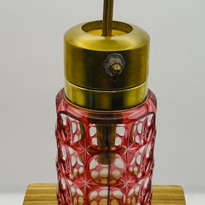 May include: A vintage perfume bottle with a red, patterned glass body and a brass top. The bottle has a long brass stem and a circular brass stopper. The glass has a repeating geometric design. The bottle is on a wooden surface.