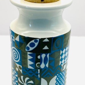 Può includere: Una bottiglia in ceramica bianca con tappo in sughero. La bottiglia presenta un motivo geometrico nei toni del blu, verde e bianco. Il tappo in sughero è marrone chiaro. L'altezza della bottiglia è di circa 20 cm.