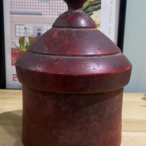 May include: A dark red, antique-style wooden container with a lid. The container is cylindrical with a slightly flared base and a domed lid topped with a small finial. The wood has a distressed finish, suggesting age and use. The container is sitting on a light-colored surface.