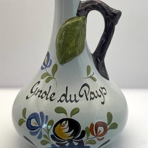 Pode incluir: Uma jarra de cerâmica azul claro com uma alça marrom e um design de folha verde. A jarra tem um padrão floral e o texto "Gnole du Pays" em escrita preta.